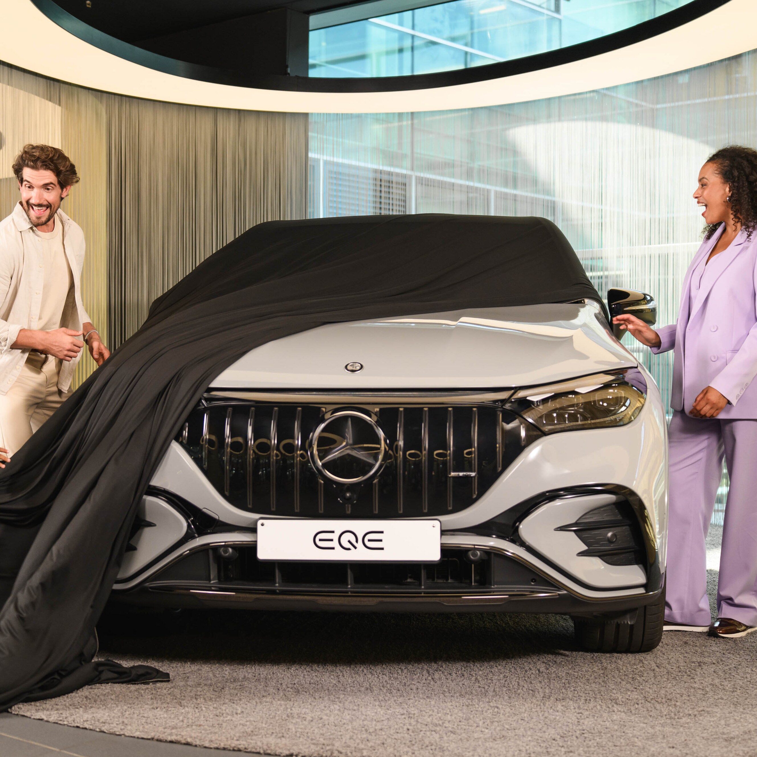 Two people and a child removing a cover off a new Mercedes-Benz vehicle in a showroom.