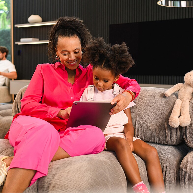 A person and child sitting on a sofa using a mobile device, with another person sitting in the distance.
