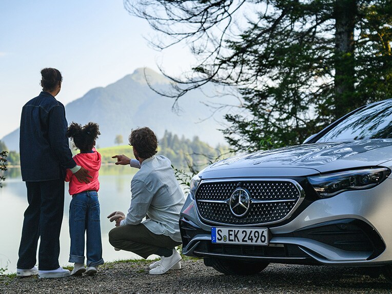 Two people and a child looking at a mountain in the distance whilst next to a Mercedes-Benz vehicle.