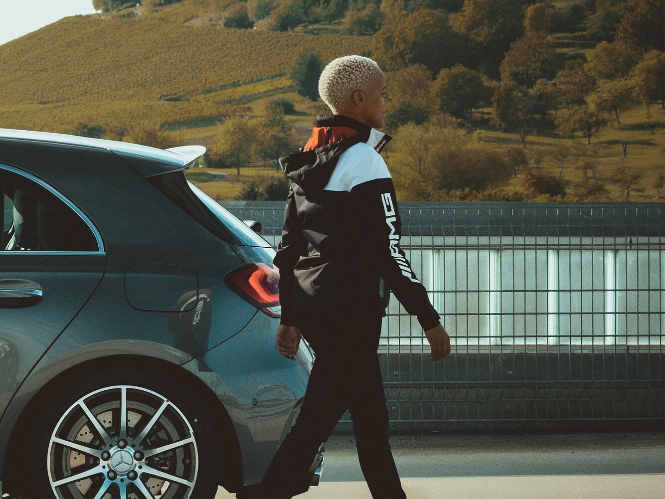 A young woman in an outfit from the AMG Racing Collection walking next to her Mercedes.